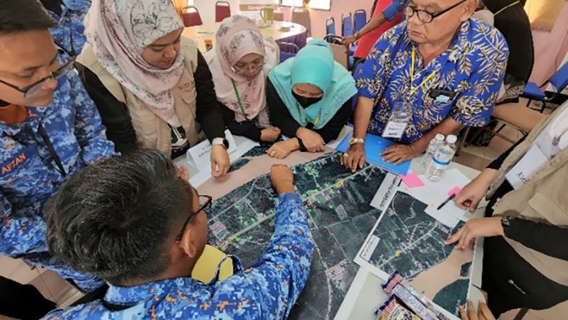 People stand grouped around a table, working together on mapping safer locations for disaster relief and emergency rescue 
